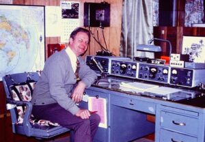 Frank Darmofalski smiles warmly, seated at a steel desk that is his home ham radio station.